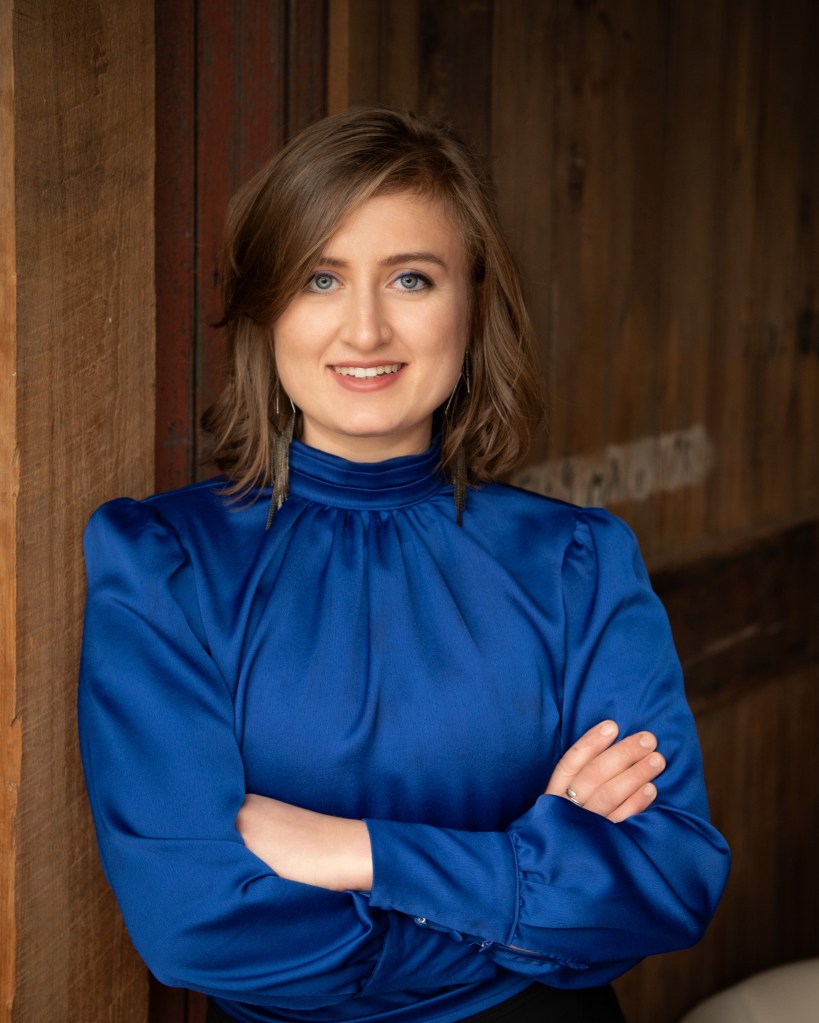 Alex Schanne standing in a power pose in a blue satin blouse.